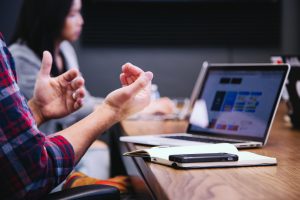 Richtig digital kommunizieren heute im Unternehmen. Auf dem Tisch steht ein Laptop, sowie Notizbuch und Handy. Davor die Hände eines Mitarbeiters, dessen Gestik eine Konversation andeutet. Die wichtigsten Kommunikationsskills 2019 