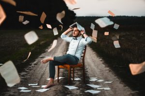 Mann sitzt auf dem Stuhl in der Natur und über ihm fliegen Bücher. 